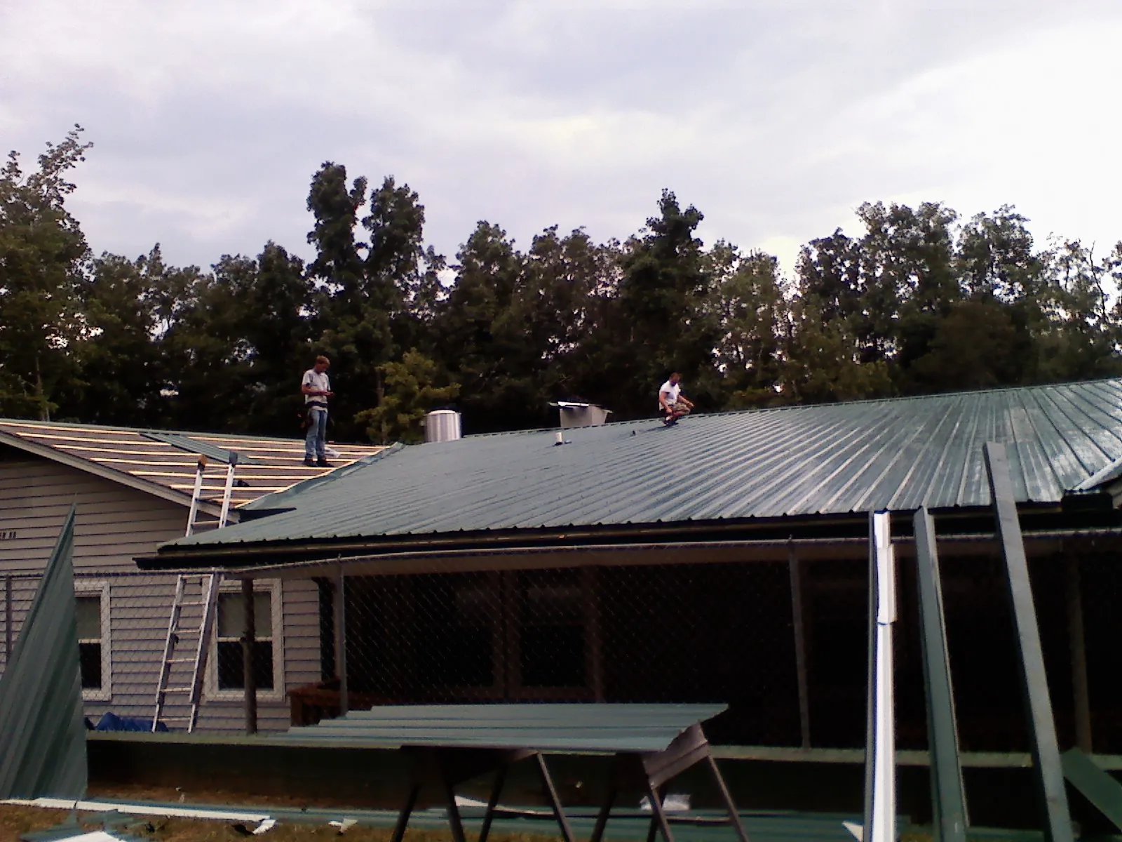 Two roofers installing green metal panels above a covered porch in Petersburg